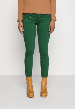 Personne portant un pantalon slim vert, un haut à manches longues couleur camel et des bottines beige, se tenant sur une surface plane.