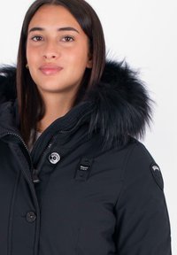 Zwarte parka jas met een nep bont capuchon, rits- en knoopsluitingen, voorzien van schouderpatches en merkaccenten op de borst.