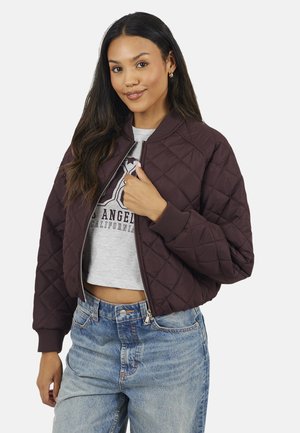 Femme portant une veste matelassée sombre par-dessus un t-shirt court à motif et un jean bleu, souriante avec les cheveux foncés lâchés.