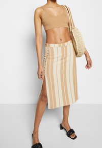 Kvinna i beige ribbad crop top, beige och krämfärgad midikjol med lodräta ränder och sidoklyfta, svarta klacksandaler och bärandes en vävd toteväska.