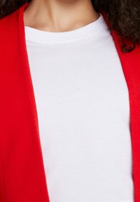 Rode cardigan over een witte ronde hals t-shirt, met een zachte textuur en een recht gesneden ontwerp. Focus op levendige kleur en gelaagd uiterlijk.