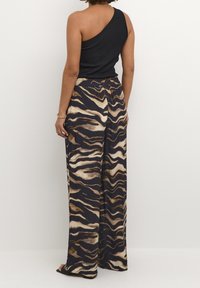 Nainen, joka käyttää leveälahkeisia eläinkuosilla varustettuja housuja ja mustaa olkaimetonta hupparia, seisoo tasaisen valkoisen taustan edessä.