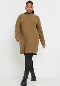 Braunes Strickkleid mit einem Reißverschlusskragen, langen Ärmeln und lockerer Passform, kombiniert mit schwarzen Strumpfhosen und klobigen Absatzstiefeln.