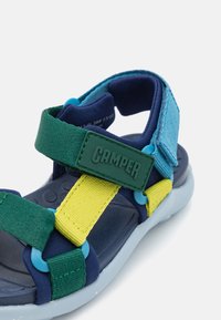 Flerfärgad sandal med gröna, marinblå och gula remmar, blå accenter och en texturerad fotbädd. Inkluderar en grön logotagg.