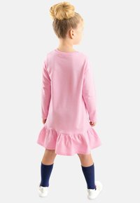 Vaaleanpunainen pitkähihainen mekko, jossa on röyhelöhelma, valmistettu pehmeästä materiaalista. Yhdistetty tummansinisiin polvihousuihin ja valkoisiin tenepalloihin.