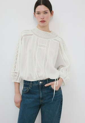 Frau trägt eine weiße Bluse mit Spitzenverzierung, drei Viertel Ärmel und blaue High-Waist-Jeans, stehend vor einem schlichten weißen Hintergrund.