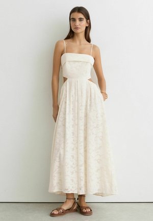 Jeune femme portant une robe en dentelle florale blanche sans manches avec des découpes sur les côtés et des sandales plates marron, debout devant un mur blanc uni.