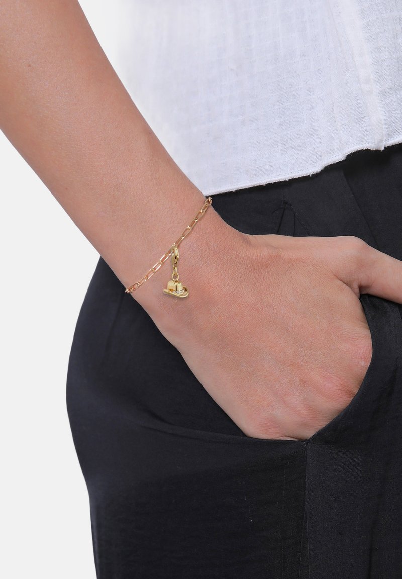 Guldkedjearmband med en liten hjärtdetalj, bärs på handleden, med stora länkar och polerad finish. Armbandet kompletterar en svart outfit.