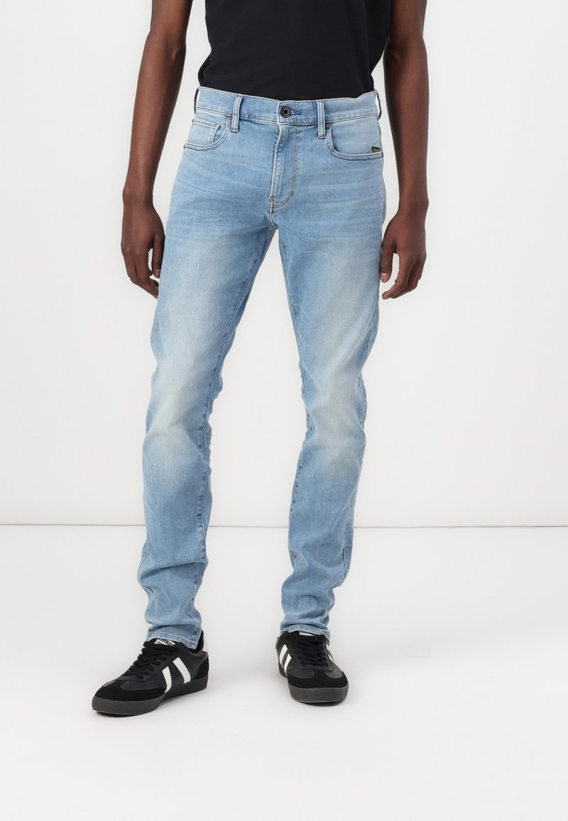 Jeans de corte ajustado de color azul claro, usados por una persona con zapatillas negras y una camiseta negra, de pie frente a un fondo blanco.