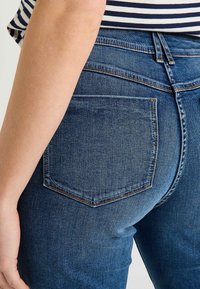 Jean en denim bleu foncé avec une coupe ajustée, dotée d'une poche arrière unique, de coutures discrètes et d'une texture lisse.