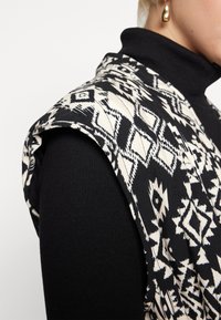 Svart och vit ärmlös jacka med ett geometriskt mönster, tillverkad av mjukt tyg, lager-på-lager över en svart halsduk, med en strukturerad axel.