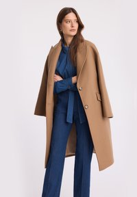 Kobieta z długimi brązowymi włosami, nosząca złote kolczyki, niebieski dżinsowy kombinezon oraz beżowy płaszcz zarzucony na ramiona, patrzy w prawo.