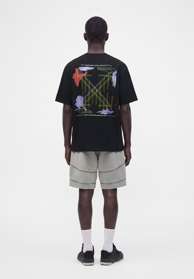 Άνδρας που φοράει μαύρο γραφικό T-shirt με πολύχρωμες αφηρημένες γραμμές και σχήματα στην πλάτη, γκρι σορτς, λευκές κάλτσες και μαύρα παπούτσια.