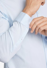 Camicia azzurra chiara con maniche lunghe, texture liscia, dettagli con bottoni sui polsini e design aderente.