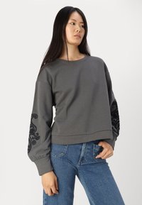 Felpa grigia con dettagli ricamati neri sulle maniche. Presenta un colletto rotondo e un design leggermente corto, abbinata a jeans in denim blu.