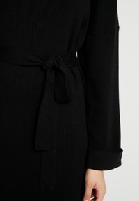 Svart ribbad klänning med långa ärmar, en knytbar midjebälte och rullad ärmdetalj för en strömlinjeformad silhuett. Mjuk textur och mångsidig design.