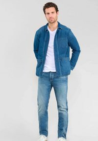 Veste en jean bleu foncé avec un col classique, fermeture à boutons et poche poitrine, assortie avec un jean bleu clair et des baskets blanches.