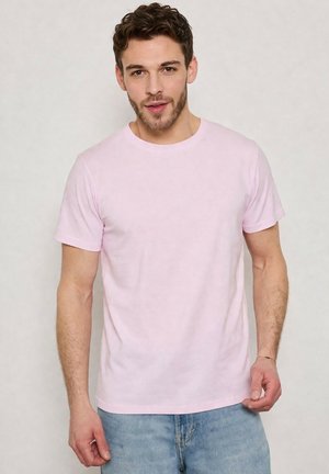 Homme aux cheveux courts et bouclés et à la barbe, portant un t-shirt rose clair à manches courtes et un jean bleu, debout devant un fond clair uni.