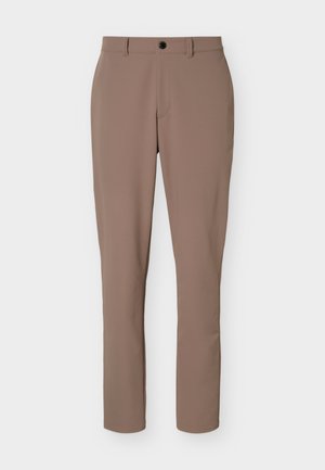 Hombre de pie con una camiseta negra de manga larga con cremallera, pantalones beige con cinturón negro y zapatillas blancas frente a un fondo claro y liso.