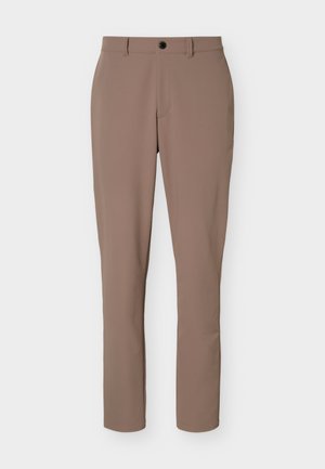 Hombre de pie con una camiseta negra de manga larga con cremallera, pantalones beige con cinturón negro y zapatillas blancas frente a un fondo claro y liso.