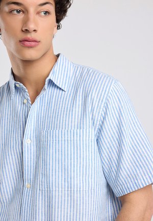 Junger Mann trägt ein blau-weiß vertikal gestreiftes Kurzarmhemd mit Knöpfen und schaut seitlich vor einem einfarbigen Hintergrund.
