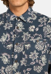 Bloemenpatroon overhemd met knopen in donker marineblauw, met groene en roze bladeren, een puntige kraag en bruine knopen. Het heeft een borstzak.