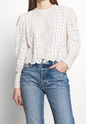 Hvid langærmet bluse med hulmønster i blonder og pufskuldre, kombineret med højtaljede lyseblå jeans, den ene hånd i lommen.