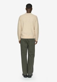 Person iført en beige sweater, olivengrønne bukser og sorte sko, stående med ryggen til en ensfarvet hvid baggrund.