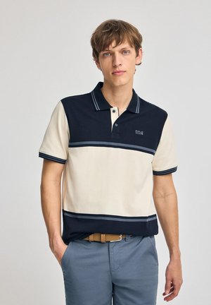 Polo-shirt i navy og creme, lavet af bomuld. Har en to-knaps placket, kontrasterende striber og et logo på brystet. Korte ærmer.