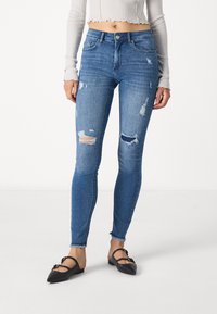Skinny blue denim jeans with distressed rips and frayed hems. Features traditional five-pocket design and brass button closure.