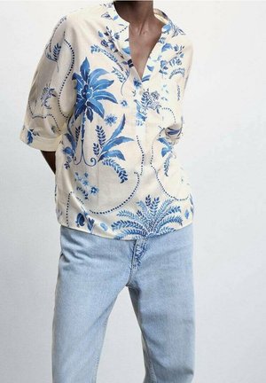 Person iført en cremefarvet bluse med blå blomsterprint og lyseblå jeans, stående mod en ensfarvet, lysegrå baggrund.