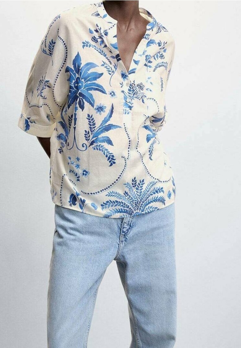 Personne portant un chemisier crème avec des motifs floraux bleus et un jean bleu clair, debout devant un fond uni gris clair.