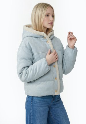 Frau trägt hellblaue Steppjacke mit Sherpa-Futter und Kapuze, befestigt Holzknöpfe, kombiniert mit blauen Jeans, vor weißem Hintergrund.