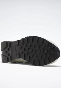 Svart gummisula med upphöjda cirkulära mönster och texturerade kanter. Ljusfärgad övre sektion övergår i sulan. Logotypen är synlig.