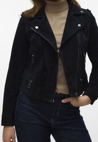 Černá semišová motocyklová bunda se stříbrným kováním, předním zipem a několika zipovými kapsami. Stylizováno s béžovým rolákem a tmavými džíny.