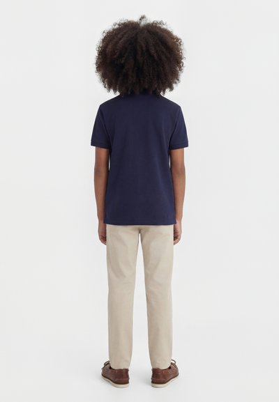 Hullámos hajú gyerek, tengerészkék rövid ujjú galléros pólót, bézs nadrágot és barna cipőt visel, háttal áll fehér háttér előtt.