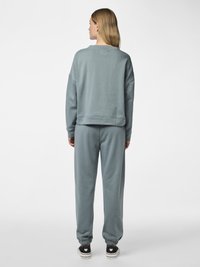 Kvinne med langt blondt hår sett bakfra, iført en løs grå sweatshirt og matchende joggebukser, med svarte joggesko på ensfarget bakgrunn.