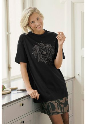 Schwarzes oversized T-Shirt mit kurzen Ärmeln, ausgestattet mit einem runden silbernen Verzierungsdesign. Kombiniert mit einem gemusterten khakifarbenen Rock.