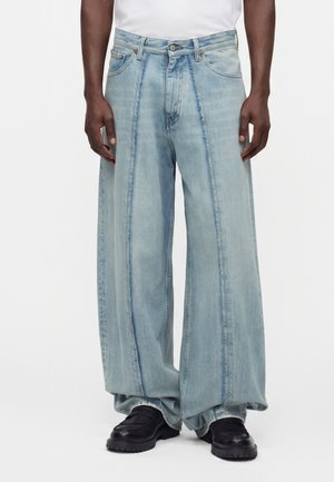 Mand iført lyseblå vidde jeans med lodrette sømdetaljer, en hvid skjorte og sorte chunky sko på hvid baggrund.