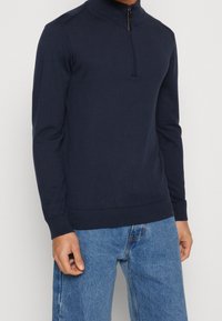 Uomo che indossa un maglione blu navy a maniche lunghe con zip a quarto e jeans in denim azzurro, in piedi con le braccia rilassate lungo i fianchi.