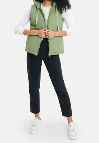 Veste matelassée verte avec capuche, fermeture éclair et cordons, portée sur une chemise blanche à manches longues, associée à un jean noir et des baskets blanches.