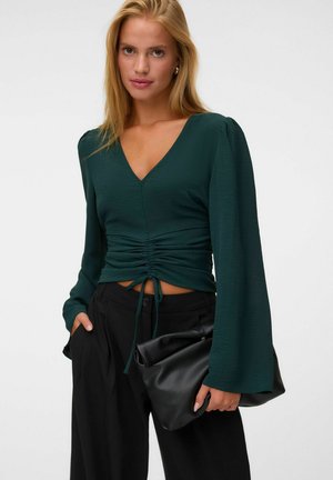Jeune femme aux cheveux blonds et raides porte un top froncé vert foncé à manches longues et un pantalon noir, tenant une pochette en cuir noire pliée.