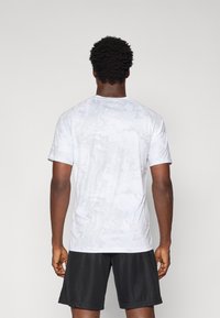 Hvid kortærmet træningsskjorte med et subtilt camouflage-mønster, lavet af letvægtsmateriale. Bæres sammen med sorte shorts.