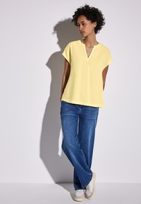 Maglietta gialla a maniche corte con scollatura a V, abbinata a jeans a gamba larga blu e sneakers bianche. Tessuto liscio con una vestibilità comoda.