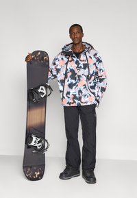 Snowboard con grafica marmorizzata nera, bianca e arancione. Il modello indossa una giacca con cappuccio multicolore e pantaloni e stivali impermeabili neri.