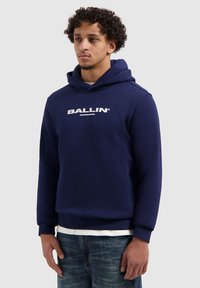 Donkerblauwe hoodie met een wit "BALLIN"-logo op de voorkant, voorzien van een capuchon met trekkoord en geribbelde manchetten, gecombineerd met een blauwe spijkerbroek.