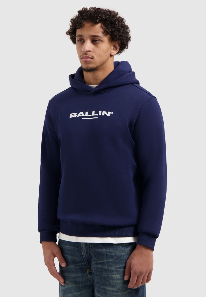 Donkerblauwe hoodie met een wit "BALLIN"-logo op de voorkant, voorzien van een capuchon met trekkoord en geribbelde manchetten, gecombineerd met een blauwe spijkerbroek.