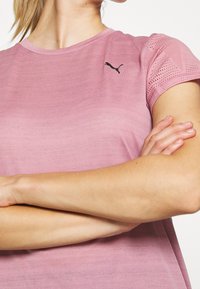 Woman crossing arms wearing a pink Puma short-sleeve athletic shirt with mesh detailing on the sleeves and black Puma logo on chest.