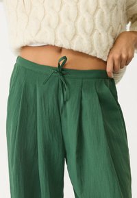 Persoon in hoge groene broek met een gestrikt tailleband en een cropped crèmekleurig kabelgebreid sweater, hand rustend nabij de zak.