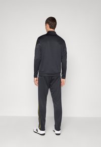 Zwart sportief trainingspak met een aansluitend ontwerp, voorzien van gele zijkanten op de broek en een gladde textuur. Het komt met lange mouwen en een kraag.